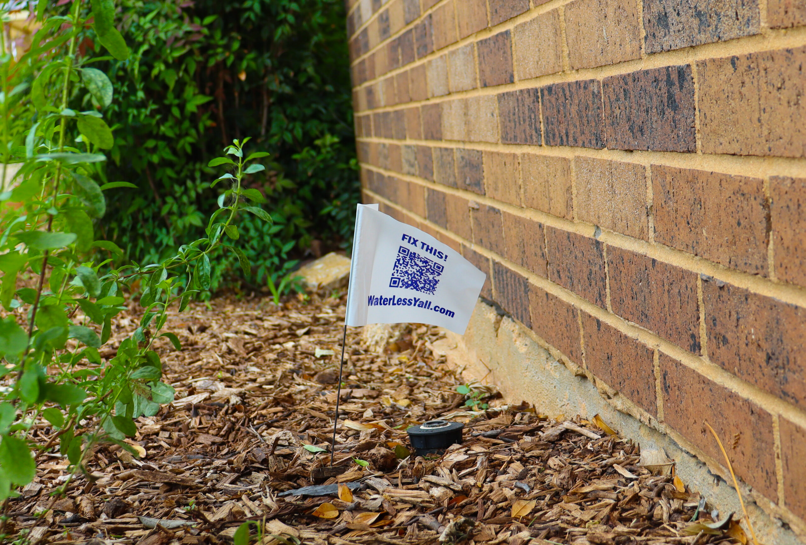 Sign Up for Irrigation Checkup 2 - UTRWD Small flag on a stake planted in mulch beside a brick wall, showing the text 'FIX THIS' and a QR code.
