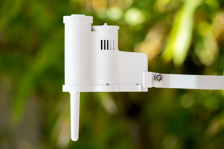 Close-up of a white motor or actuator housing mounted on a metal arm outdoors, with a vertical antenna-like projection and a blurred green background.