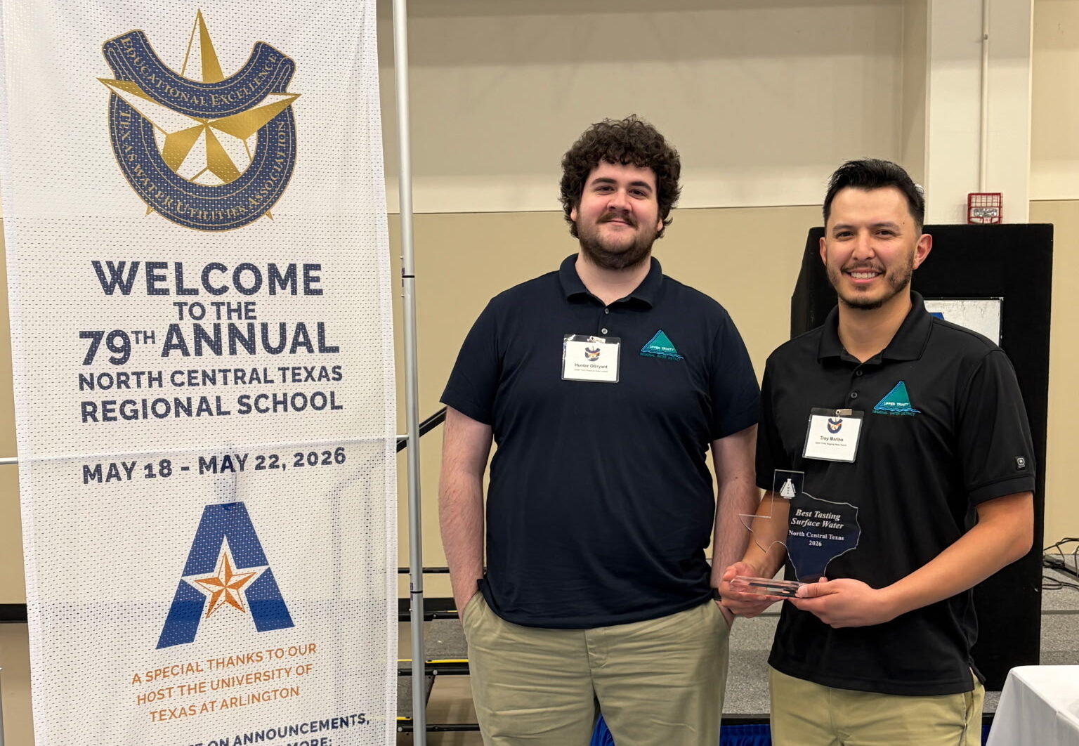 Two men in polo shirts with name tags stand beside a tall banner for the 79th Annual North Central Texas Regional School, one holding a glass award at the event venue.
