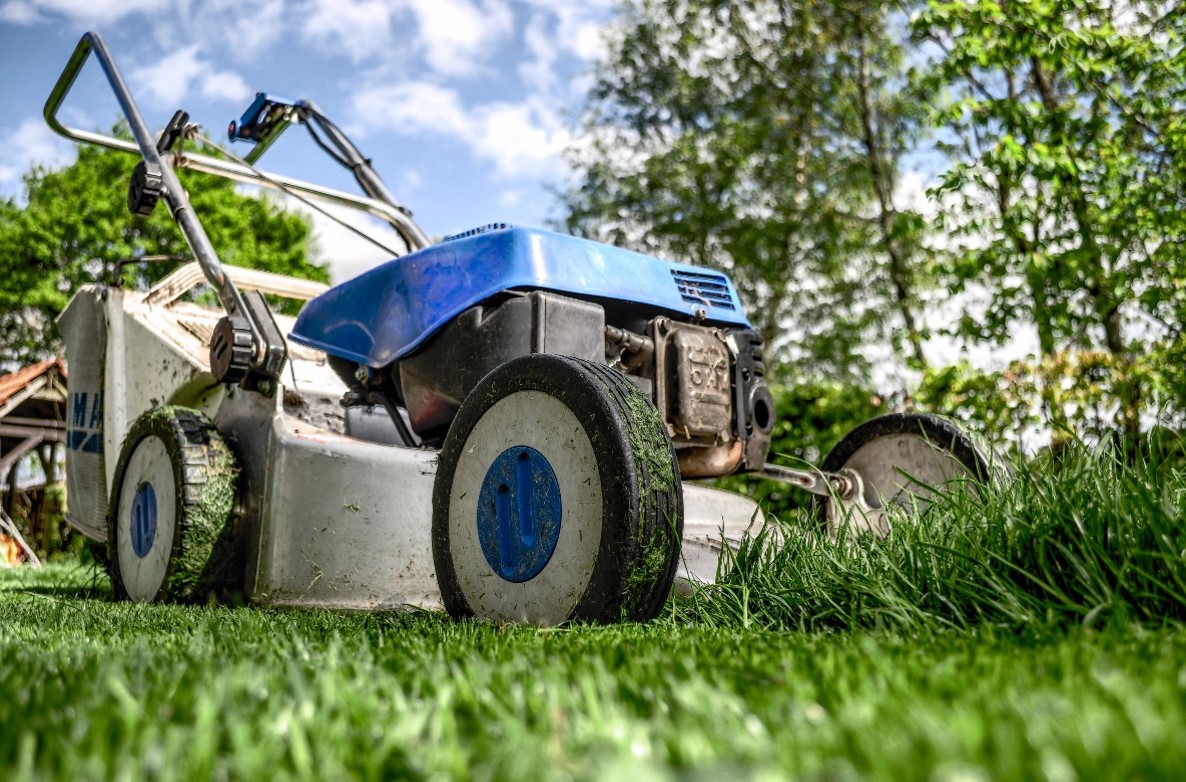 Blue and gray riding lawn mower resting on a green lawn with trees and a sunny sky in the background.