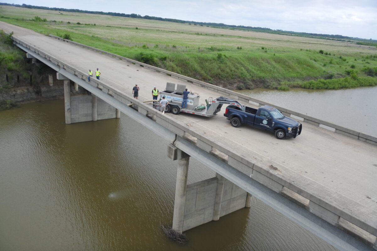 Texas Parks and Wildlife crew stand on SH 34 bridge and port fish from tank truck into the North Sulphur River channel inside the lake footprint.