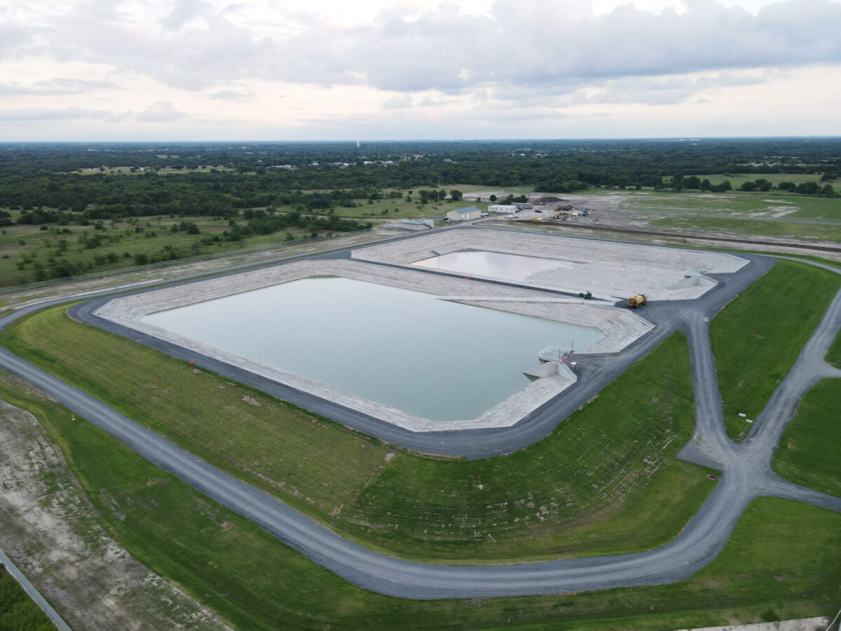 Aerial view of a large rectangular concrete water basin surrounded by roads and green fields, with a construction yard in the distance.